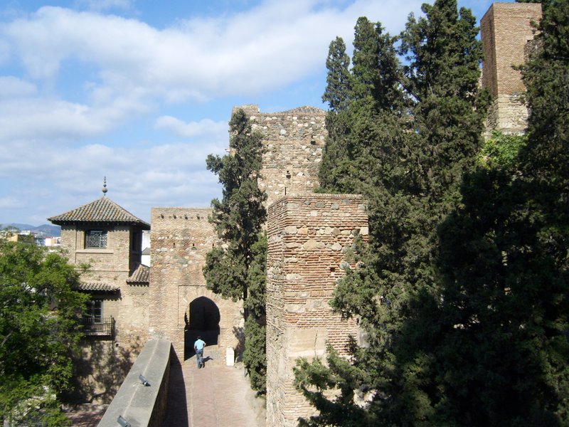 Blick vom Wehrgang der maurischen Festung Alcazaba in Málaga, Spanien, in Richtung eines Torbogens und eines Wehrturms, umgeben von hohen Zypressen und typischen Ziegelmauern aus der Nasridenzeit